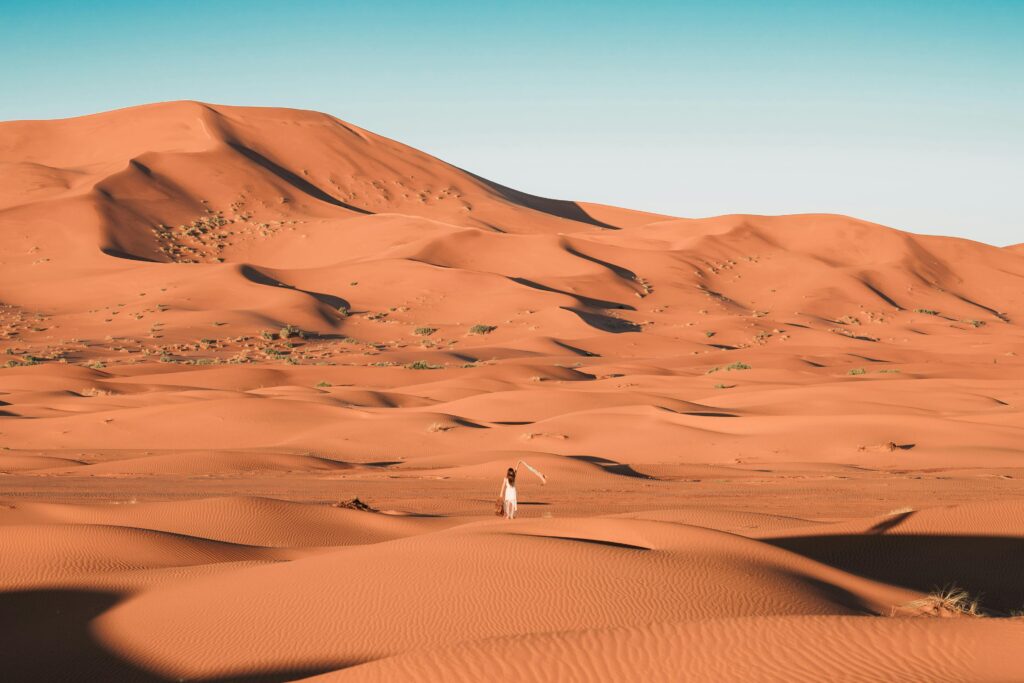 A lone person walking amidst the vast sand dunes of the Sahara Desert under a clear blue sky.