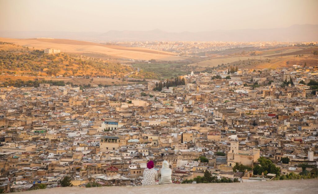 Back view of unrecognizable distant female in traditional wear and headwear sitting on top of mountain above old town with buildings